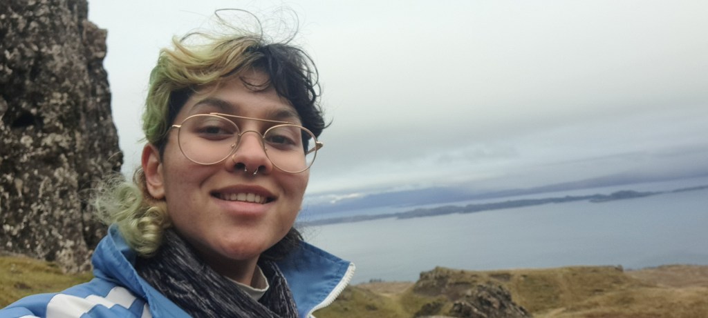 Landscape image of Sabrina smiling at the camera, wearing a blue zip jacket. They are a person with half green and half black split hair, wearing glasses, standing in front of a rock in front of the sea.